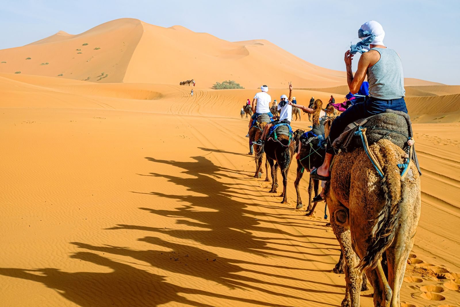 A group of people riding camels across the vast sand dunes under a bright sky.